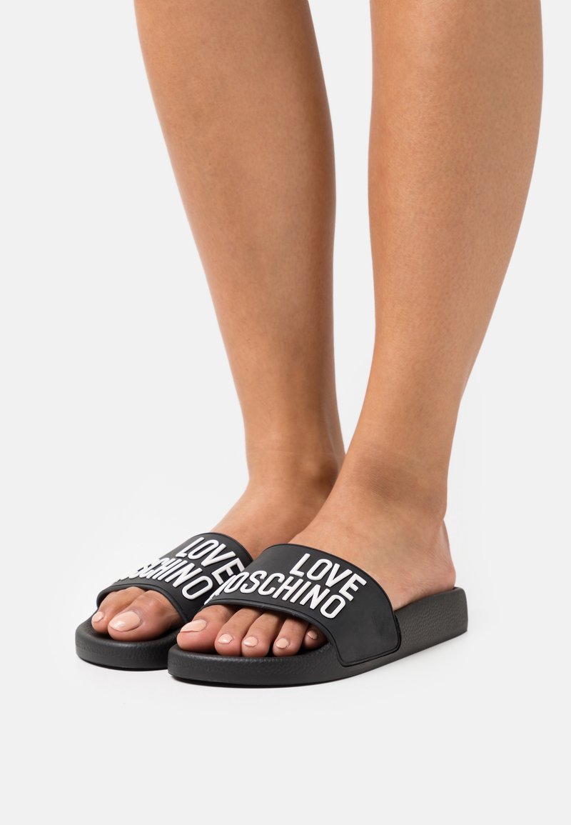 Pieds portant des sandales noires sans bride avec un texte blanc "LOVE MOSCHINO" sur les brides sur un fond clair uni.