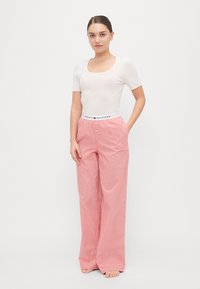 Pantalons larges rose pâle en tissu léger, avec une ceinture élastique blanche portant un logo. Le modèle porte un haut blanc ajusté.