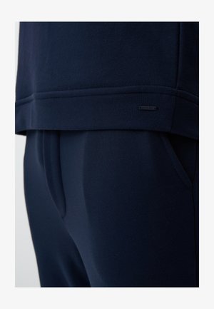 Dunkelblaues Loungewear-Set aus einem weichen, strukturierten Oberteil und dazu passenden Hosen, mit einem dezenten Logo-Patch am Saum.