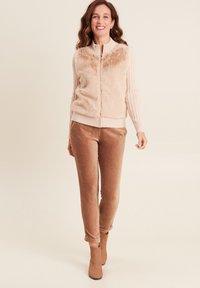 Sweater beige en fausse fourrure avec fermeture éclair et manches côtelées, associé à un pantalon marron en peluche, coupé fuselé, et des bottillons assortis. Texture lisse dans l'ensemble.