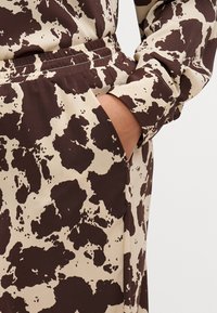 Tela crema con manchas marrones oscuras, cuenta con una cinturilla elástica y un bolsillo lateral, mostrando una textura suave y mate.