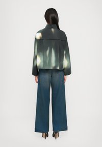 Übergroße dunkelgrüne Jacke mit glatter Textur, kombiniert mit weit geschnittenen Jeans in mittlerem Blau. Hochhackige schwarze Schuhe vervollständigen den Look.