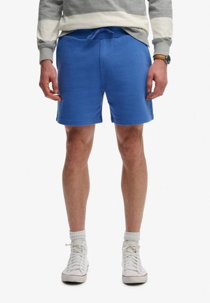 Hombre de pie con pantalones cortos azules con cordón, zapatillas blancas altas, calcetines blancos y una camiseta de manga larga gris y blanca con reloj.