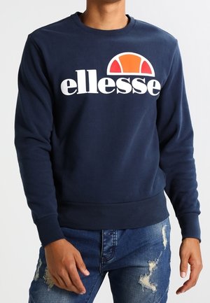 Mand iført marineblå sweatshirt med hvidt "ellesse" logo og orangerødt halvcirkeldesign, kombineret med slidte blå jeans.