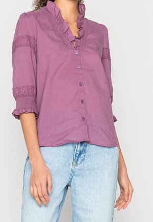 Femme portant un chemisier violet à boutons avec col à volants et bordure en dentelle aux manches, associé à un jean bleu clair.