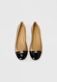 Ballerine in pelle color cuoio con punta in patent leather nera e dettaglio logo dorato sul davanti. Texture liscia, forma arrotondata.