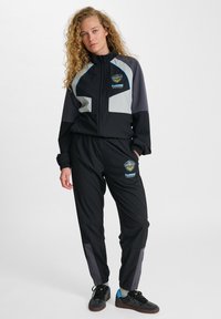 Svart träningsoverall med grå och blå accenter, bestående av en jacka med dragkedja och byxor med elastisk midja. Logotypen syns på bröstet och benet.