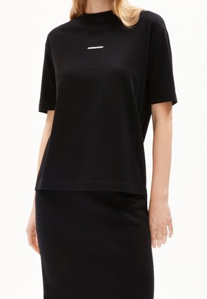 Femme portant un haut à manches courtes noir ample et une jupe noire, avec un petit texte blanc "ARMEDANGELS" centré sur la poitrine.