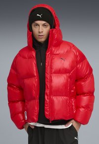 Rode pufferjack met een glanzende textuur, voorzien van een capuchon, rits aan de voorkant en logodetail. Combinatie met een zwarte hoodie en donkere broek.
