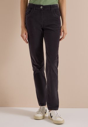 Personne portant un pantalon droit de couleur foncée, une chemise verte et des baskets blanches avec motif noir, debout sur un fond beige.