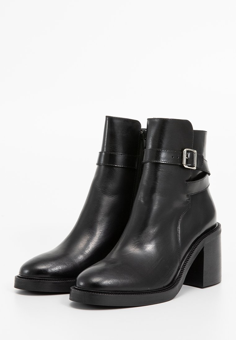 Stivali neri in pelle alla caviglia con tacco spesso, dotati di una cerniera laterale e un dettaglio a fibbia sulla fascia attorno alla caviglia. Texture liscia.