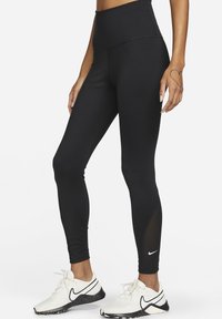 Osoba ubrana w czarne legginsy z wysokim stanem i białe sportowe buty z czarnym logo Nike, stojąca na białym tle.