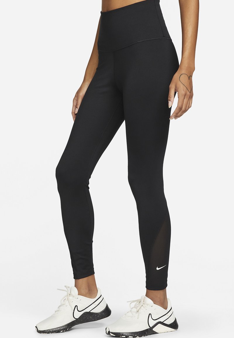 Osoba ubrana w czarne legginsy z wysokim stanem i białe sportowe buty z czarnym logo Nike, stojąca na białym tle.