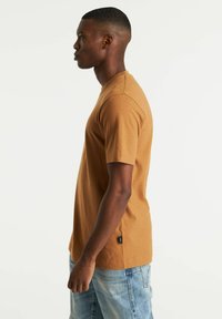 Braunes Baumwoll-T-Shirt mit kurzen Ärmeln, Rundhalsausschnitt, seitlichem Schlitz und einem kleinen schwarzen Etikett am Saum, kombiniert mit hellblauen Jeans.