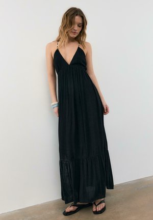 Donna che indossa un abito lungo nero senza maniche con spalline sottili, sandali e braccialetti impilati, in piedi contro un muro bianco semplice.