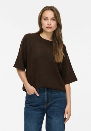 Kvinde med langt brunt hår iført en løs, mørkebrun kortærmet sweater og blå jeans, stående med den ene hånd i lommen.
