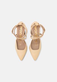 Béžové lodičky s špičatou špičkou a kotníkovými pásky, vyrobené z hladkého materiálu. Zlatý potisk loga na stélce, s elegantním designem.