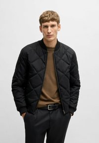 Black quilted bomber jacket with ribbed cuffs and collar, zip closure, paired with a brown textured shirt and black trousers.