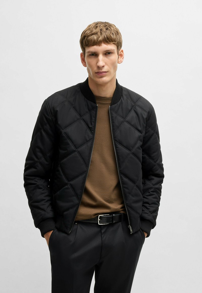 Black quilted bomber jacket with ribbed cuffs and collar, zip closure, paired with a brown textured shirt and black trousers.