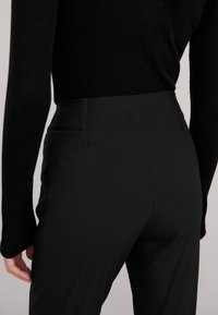 Person iført svarte høytlivs bukser og en svart langermet topp, vist fra midjen til midten av låret, stående mot en ensfarget bakgrunn.