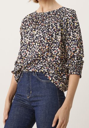 Vrouw draagt een multikleurige blouse met stippen, met een geplooide halslijn en mouwen, gecombineerd met een donkerblauwe jeans met hoge taille.