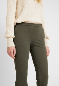Olijfgroene, stretchstof broek met een strakke pasvorm. Gecombineerd met een lichtbeige, gemêleerde gebreide trui. Geen zichtbare sluitingdetails.