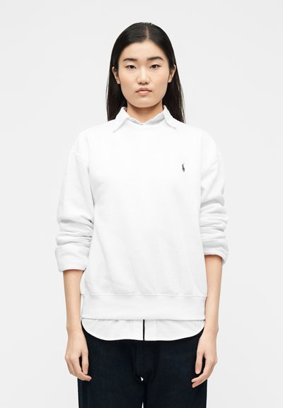 Νέα γυναίκα με ίσια σκούρα μαλλιά, φοράει λευκό φούτερ πάνω από λευκό πουκάμισο με γιακά και μαύρο παντελόνι, στέκεται μπροστά σε απλό φόντο.