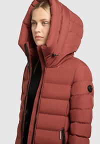 Veste matelassée rouge avec une grande capuche, fermeture éclair et poches latérales. Texture lisse, avec un logo sur la manche et des cordons ajustables.