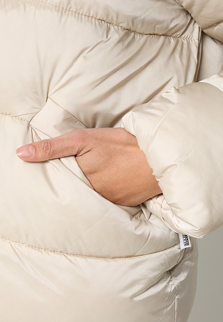 Ręka wsunięta do kieszeni jasnobeżowej kurtki puchowej z pikowanym wzorem i widocznym przeszyciem.
