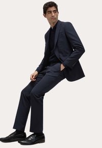 Granatowy garnitur z dopasowaną marynarką i pasującymi spodniami, połączony z czarnym polo i czarnymi loafersami. Gładki materiał z eleganckim designem.