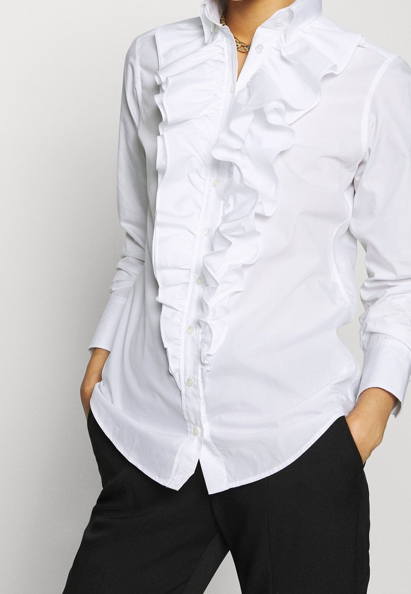 Chemise blanche à manches longues boutonnée avec un détail de volants à l'avant, portée avec un pantalon noir, mains dans les poches, sur fond clair uni.