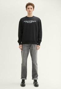 Czarna bluza z białym graficznym napisem, okrągły dekolt, luźny krój, połączona z szarymi dżinsami i czarnymi butami, stojąca na neutralnym tle.