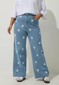 Vaaleansiniset leveälahkeiset farkut, joissa on valkoisia päivänkakkara-ompeluja ympäriinsä. Sisältää taskut ja etu-nappi sulkemisen.