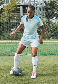 Svetlomodrý futbalový dres s krátkymi rukávmi a šortkami, doplnený o biele ponožky a topánky s červenými šnúrkami, vystavený na umelom trávniku.