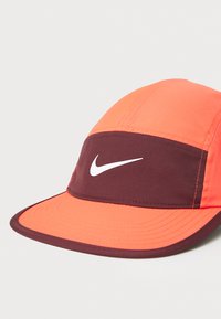 Čiapka Nike s kombináciou jasne oranžovej a burgundskej látky, s rovnou krempou a výrazným bielym logom na prednom panely.