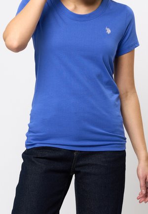 Personne portant un t-shirt bleu uni à manches courtes avec un petit logo blanc sur la poitrine et un jean bleu foncé, main près de la tête.