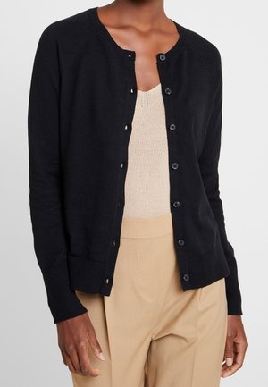 Personne portant un cardigan noir boutonné sur un top en maille beige et un pantalon beige taille haute sur un fond uni.
