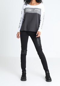 Woman wearing a black and white long-sleeve shirt with text, black patchwork pants, and black sneakers, standing against a white background.