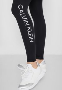 Černé legíny s bílým n�ápisem "CALVIN KLEIN" podél strany, v kombinaci s bílými síťovanými teniskami se stříbrnými akcenty a tkaničkami.