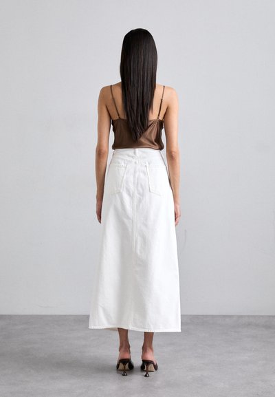 Brown satin camisole paired with a long white denim skirt featuring back pockets and seam details. Model stands on a gray floor.