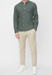 Męska lniana koszula z długim rękawem w oliwkowym kolorze, z kołnierzem stojącym, zapinana na guziki, z kieszenią na piersi; w zestawieniu z beżowymi spodniami i białymi trampkami.