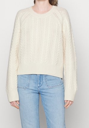 Frau trägt einen cremefarbenen, kabelgestrickten Pullover mit langen Ärmeln und hellblaue, hoch taillierte Jeans vor neutralem Hintergrund.