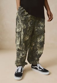 Cargo pants in a camo print featuring splattered paint patterns; made of durable fabric, with two side pockets and a relaxed fit, paired with black sneakers.