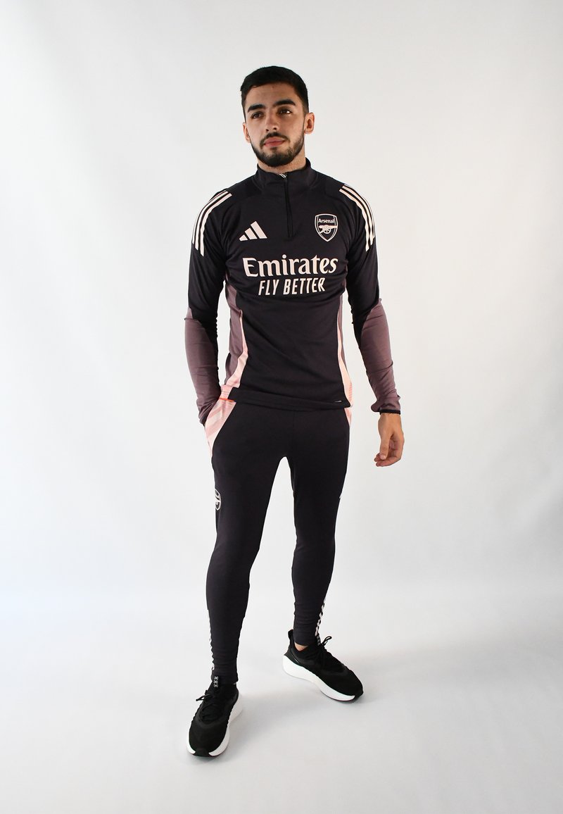Chándal deportivo negro con acentos rosas, que presenta un cuello alto y franjas de Adidas. Diseño ajustado con pantalones de longitud hasta el tobillo y detalles de logo.