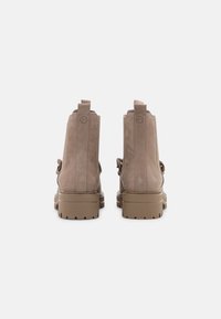 Botas de tobillo de ante marrón con una suela beige gruesa, que cuentan con una etiqueta de agarre y un detalle decorativo de correa en la parte posterior.