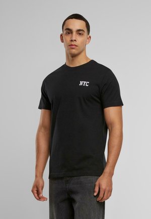 Zwarte katoenen T-shirt met korte mouwen en een ronde hals. Aan de linkerkant van de borst staat witte geborduurde tekst "NYC". Model draagt donkere jeans.