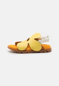 Keltaiset nahka-sandaalit, joissa on leikattuja kukkakuvioita, teksturoidut remmit ja oranssi pehmustus; ruskea kumipohja tarjoaa pitoa ja kestävyyttä.