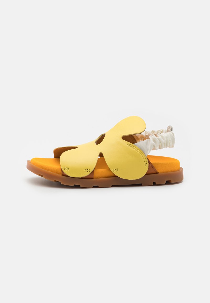 Keltaiset nahka-sandaalit, joissa on leikattuja kukkakuvioita, teksturoidut remmit ja oranssi pehmustus; ruskea kumipohja tarjoaa pitoa ja kestävyyttä.