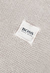 Gros plan sur un tissu beige tricoté avec une étiquette rectangulaire blanche cousue portant le texte noir "BOSS HUGO BOSS".