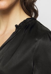 Mujer con blusa de satén negra y escote con volantes, que muestra el cuello, el hombro y parte de su cabello ondulado oscuro.